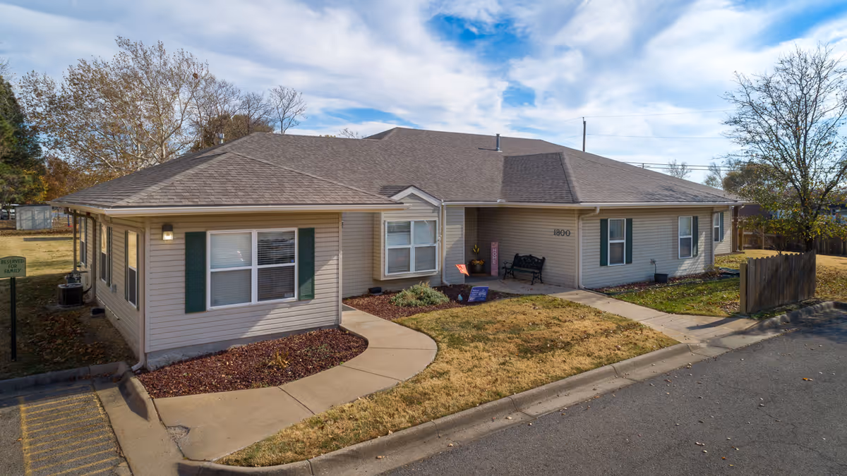 Single-story beige building with green shutters and a gray roof, surrounded by a small lawn and a sidewalk leading to the entrance. The building has the number 1800 on the front wall near the entrance, with a bench and some plants nearby. Trees and a clear blue sky with some clouds are visible in the background.