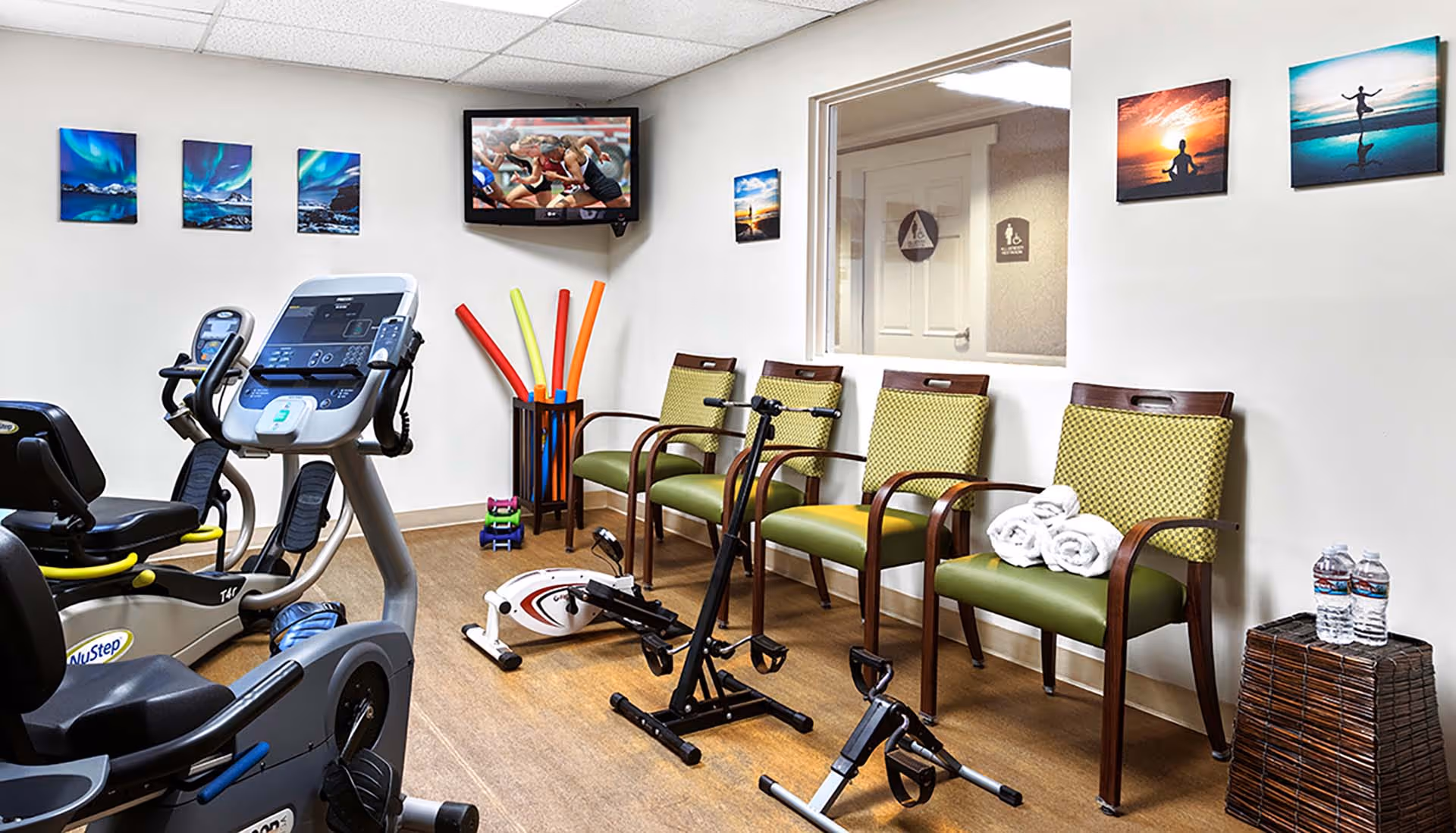 A senior living facility exercise room with stationary exercise bikes, a rowing machine, and hand pedal exercisers. Four green cushioned chairs with wooden arms are lined up against the wall, with rolled white towels on one chair and two water bottles on a small wicker table. Colorful pool noodles are stored in a corner container. Several framed pictures and a television mounted on the wall are visible.