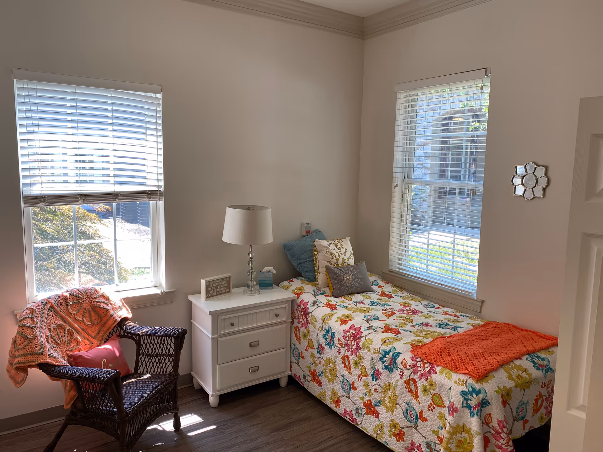 Sunlit bedroom with a single bed covered in a floral quilt next to a white nightstand and a wicker chair draped with a blanket.
