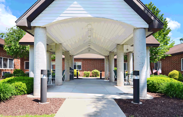 Covered outdoor patio area with a peaked roof supported by large concrete columns, surrounded by green bushes and trees, with brick buildings in the background under a blue sky with some clouds.