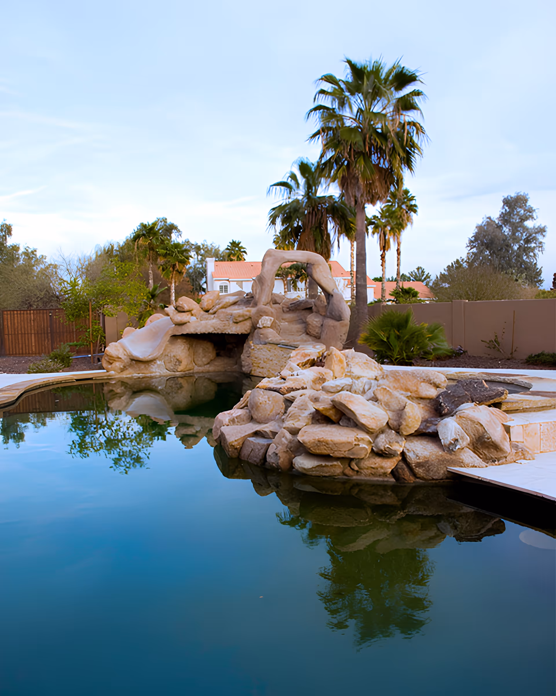 Rock-lined backyard swimming pool with a waterslide and palm trees reflected in the water.