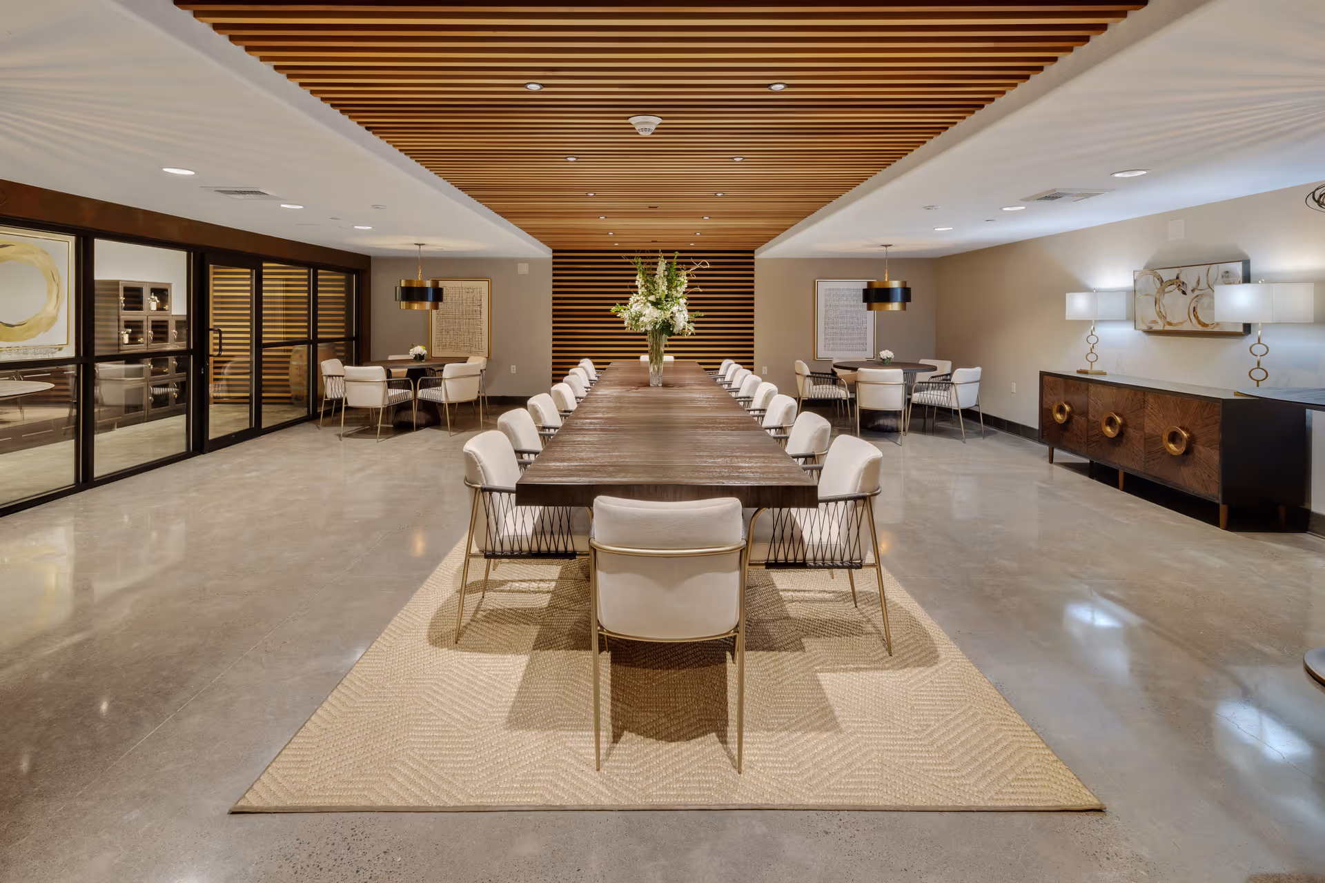A spacious, modern dining room with a long wooden table surrounded by cushioned chairs. The ceiling features wooden slats, and the floor is polished concrete with a large beige rug under the table. There are two smaller dining tables with chairs in the background, decorative wall art, and a sideboard with lamps and artwork on the right side.