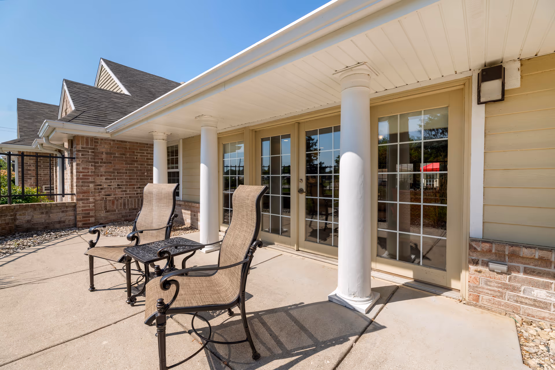 Outdoor patio area with two metal and fabric chairs and a small metal table between them, situated in front of a building with large glass double doors and white columns under a covered porch.