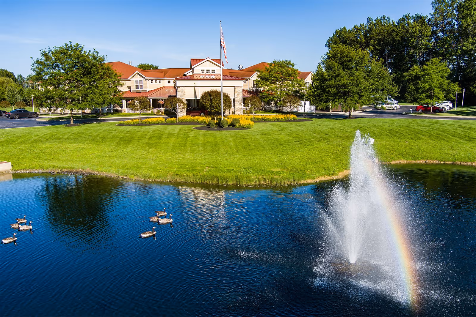 View of a senior living facility building with a red roof, surrounded by green trees and a well-maintained lawn. In the foreground, there is a pond with a water fountain creating a rainbow effect and several ducks swimming on the water.