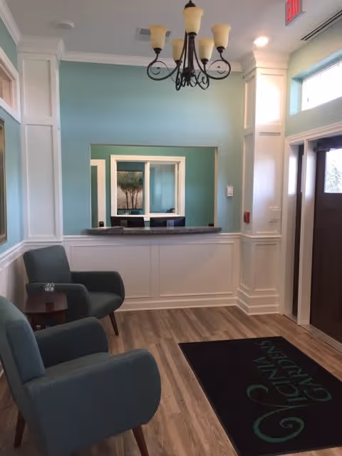 A small reception area with two teal armchairs and a small wooden side table on the left. There is a counter with a window opening behind it, framed by white and teal walls. A chandelier with five lights hangs from the ceiling. The floor is wood with a black mat that reads 'Vicinia Gardens'. To the right is a dark brown door with glass panels and a window above it, and an exit sign is visible on the ceiling.