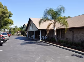 Exterior view of a single-story brick building with a covered entrance, palm trees, and parked cars along the driveway under a clear blue sky.