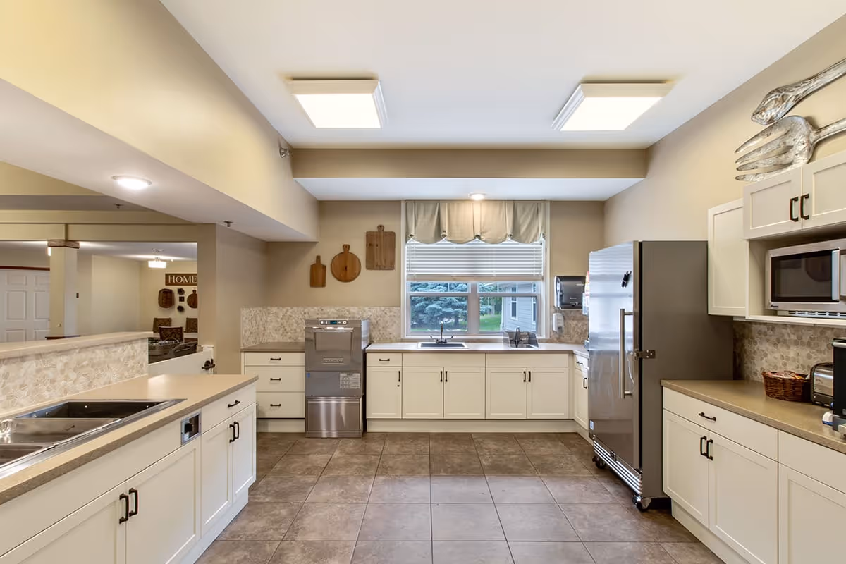 A spacious, well-lit kitchen with beige walls and tiled floor. The kitchen features white cabinets with black handles, a stainless steel refrigerator, a microwave, a dishwasher, and a double sink under a window with a valance. Decorative cutting boards hang on the wall next to the window. The kitchen opens into a living area visible in the background.