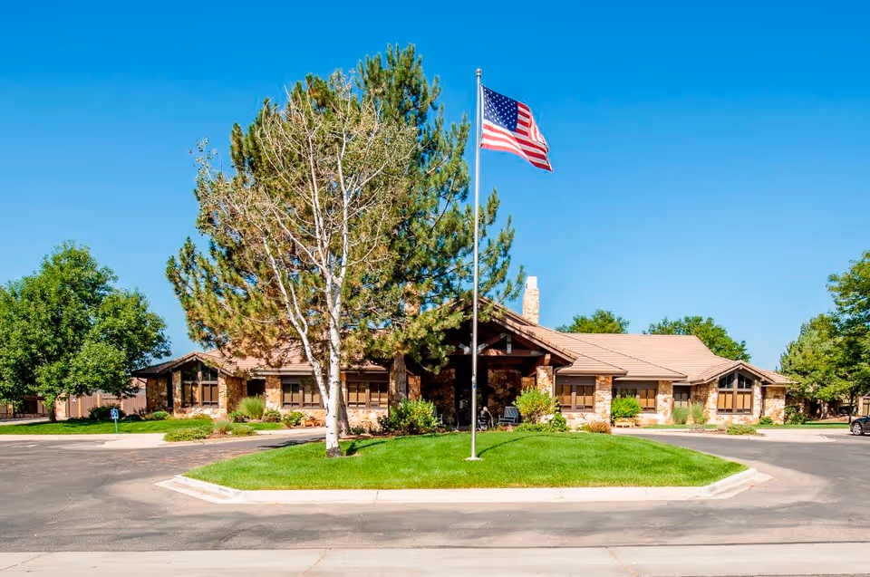 Front exterior view of a single-story assisted living facility building with stone and wood accents, surrounded by trees and greenery. An American flag is prominently displayed on a flagpole in the center of a grassy island in the driveway.