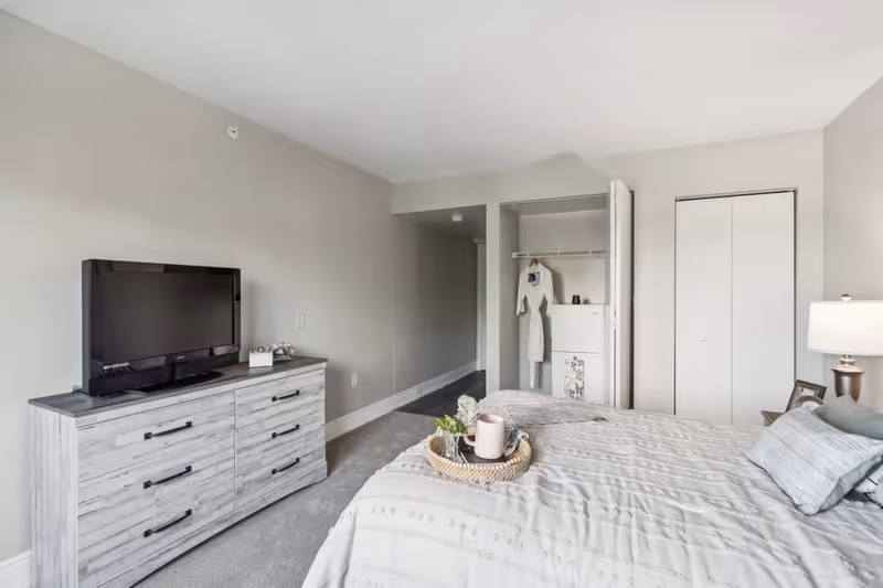 A bedroom with a bed covered in a light-colored patterned comforter and pillows. A wooden dresser with a flat-screen TV on top is positioned against the wall. There is an open closet with a white robe hanging inside and a closed closet door next to it. A lamp and framed photos are on a nightstand beside the bed. The room has light gray walls and carpeted flooring.