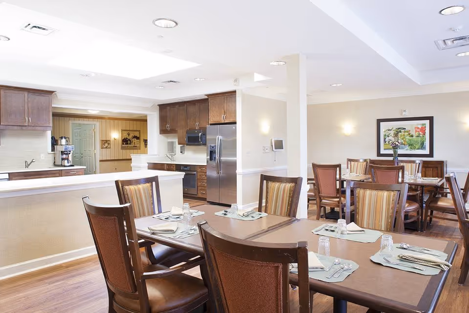 A bright dining area in a senior living facility featuring multiple wooden tables set with placemats, napkins, glasses, and silverware. The room has wooden chairs with striped cushions and a kitchen area with wooden cabinets, a stainless steel refrigerator, microwave, and coffee maker in the background. The walls are light-colored with a framed painting and soft wall lighting.