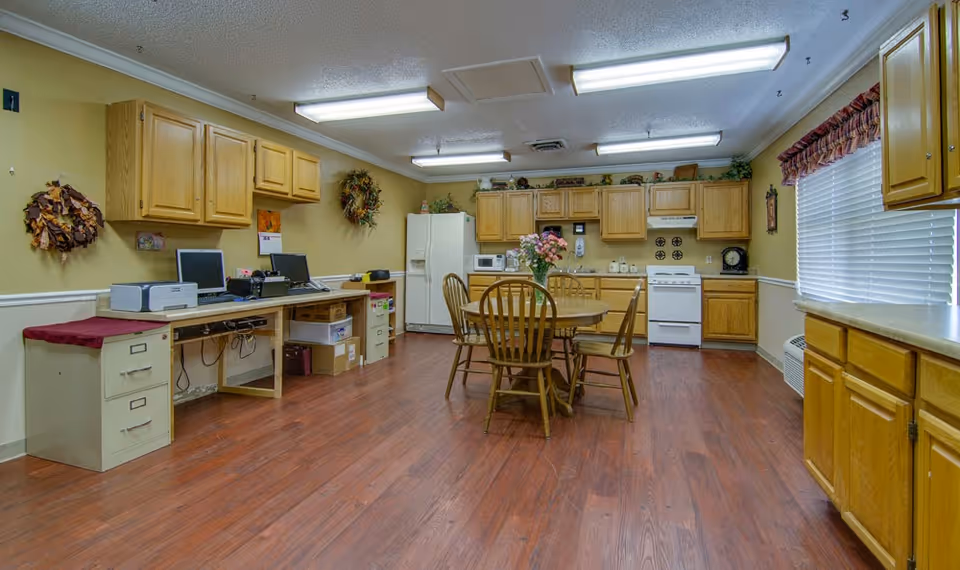 A spacious kitchen area with wooden cabinets, a white refrigerator, stove, microwave, and a round wooden dining table with four chairs. There are two computer workstations with monitors and a printer on a long desk along one wall. The floor is wooden, and the walls are painted light yellow with decorative wreaths and a calendar. A window with blinds and a valance is on the right side.