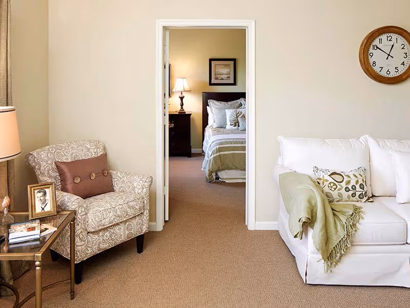 Cozy living room with an upholstered armchair, white sofa with a throw, and a view into a bedroom through an open doorway.