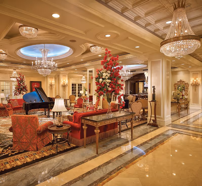 Opulent lounge with chandeliers, a grand piano, red seating, and large floral arrangements.