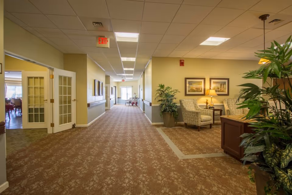 A well-lit hallway in a senior living facility with beige walls and patterned carpet. On the right side, there is a small seating area with two upholstered chairs, a side table with a lamp, framed artwork on the wall, and potted plants. On the left side, double glass doors open into a dining room with tables and chairs. The hallway extends to a bright exit door at the far end.