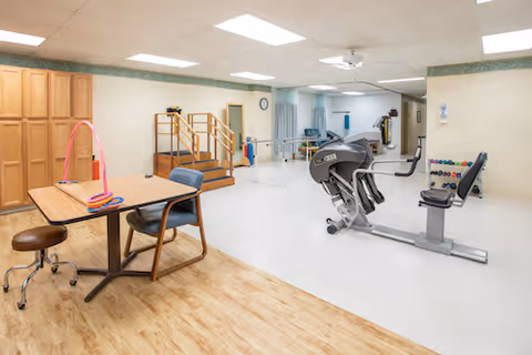 A rehabilitation therapy room with exercise equipment including a seated leg press machine, parallel bars for walking practice, a small set of stairs with handrails, a table with chairs and therapy tools, and a rack of colorful dumbbells against the wall.