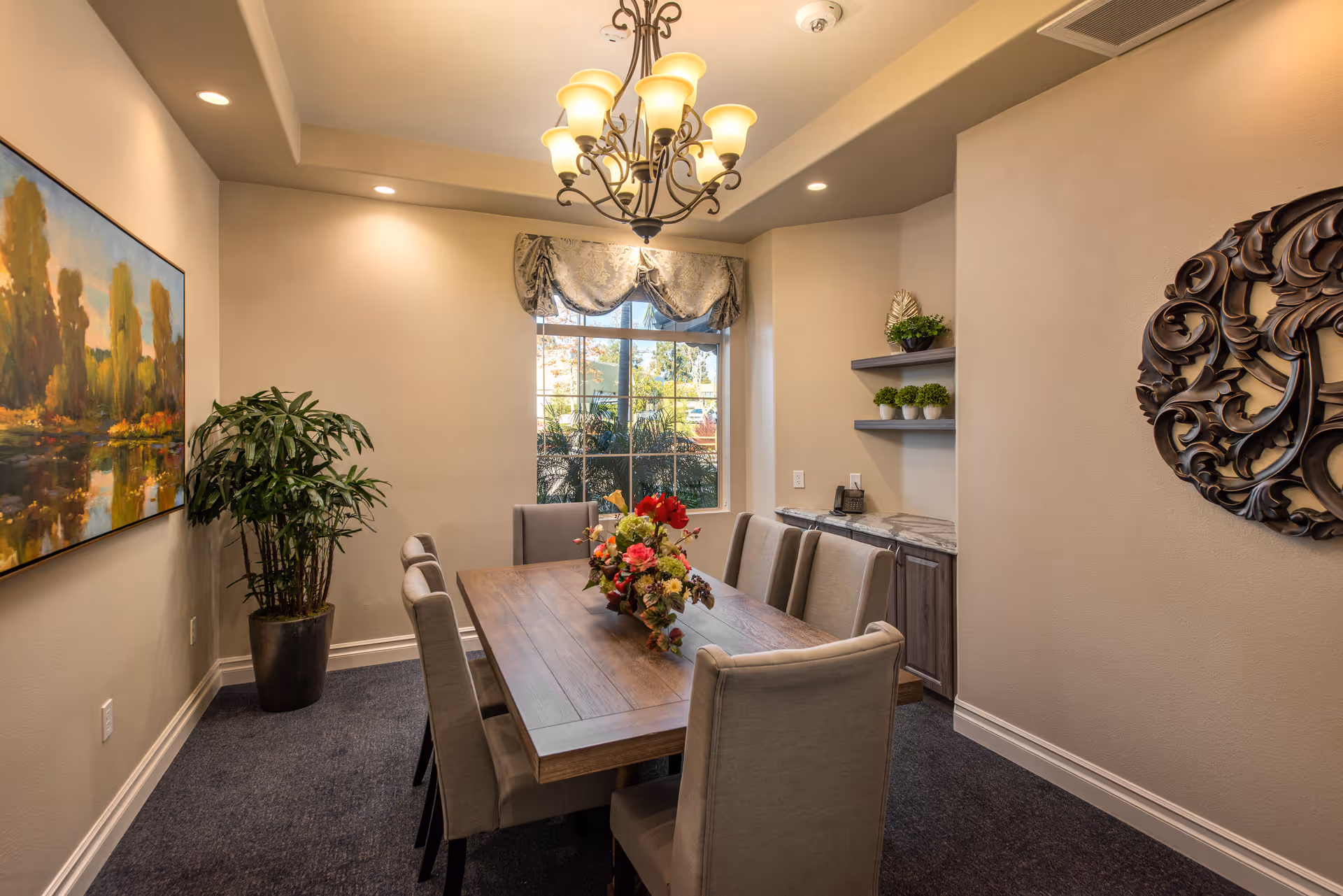 A well-lit dining room with a wooden table surrounded by eight upholstered chairs. A floral centerpiece decorates the table. There is a large window with a decorative valance, a potted plant in the corner, a landscape painting on one wall, and a decorative wooden wall art on another. Shelves with small potted plants and a countertop with cabinets are also visible.