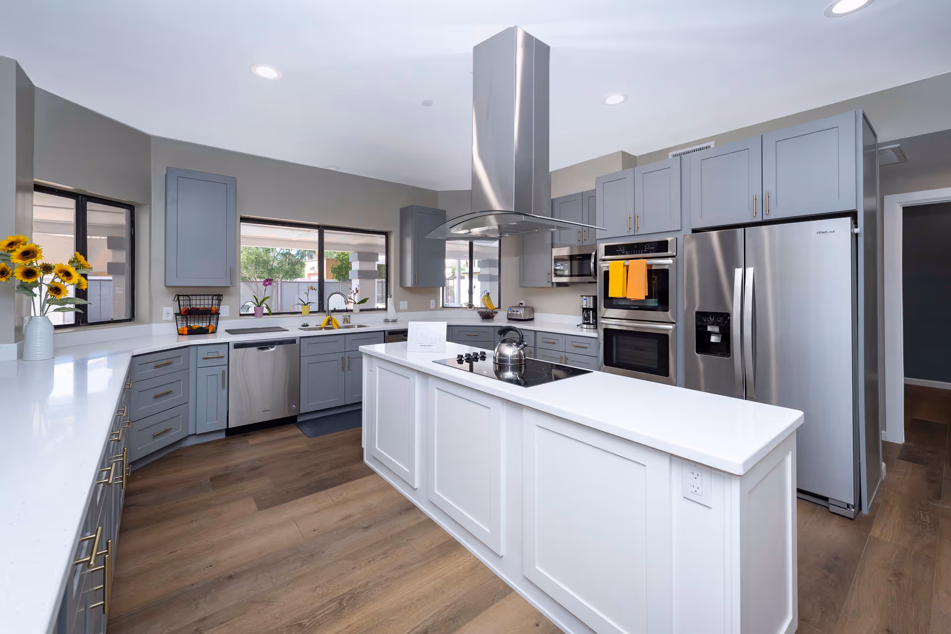 Modern kitchen with gray cabinets, white countertops, a central island with a stovetop and stainless steel range hood, stainless steel refrigerator, double oven, and dishwasher. The kitchen has large windows letting in natural light and wooden flooring.