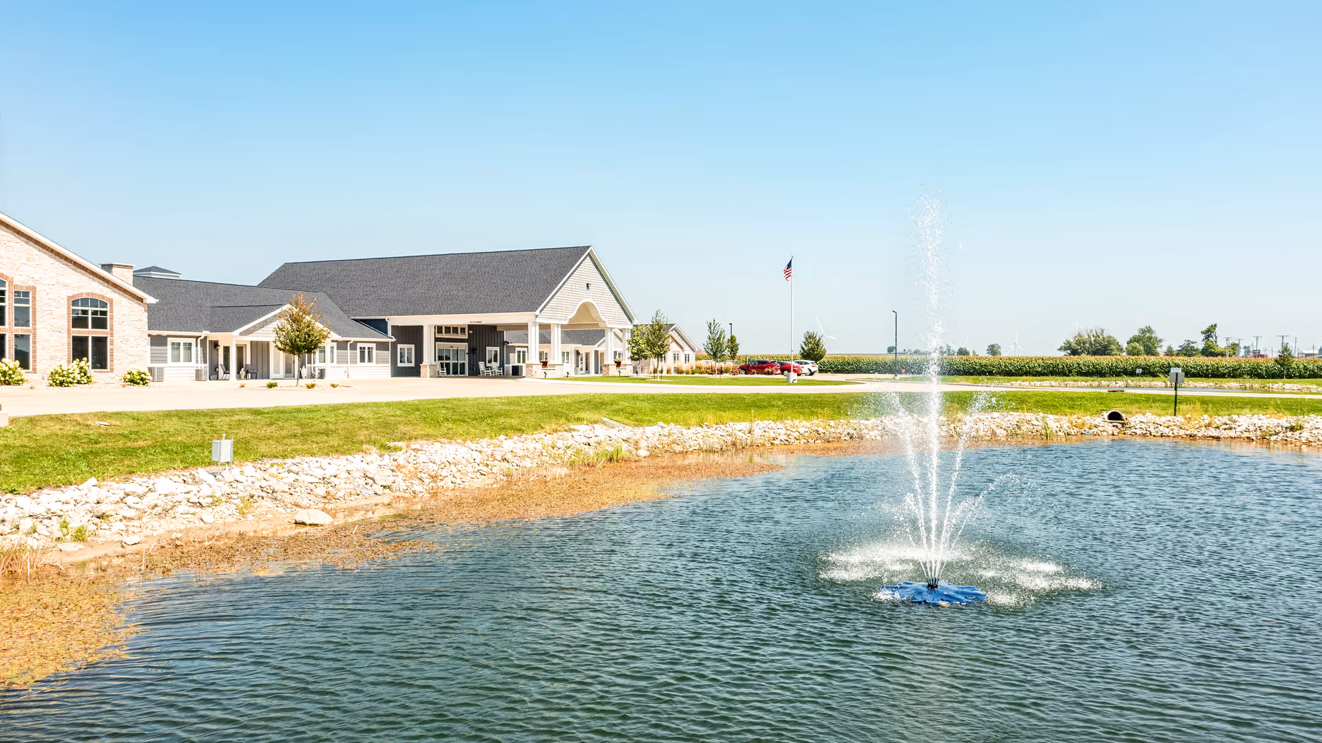 Front view of a single-story assisted living building with a pond and a fountain in the foreground under a clear sky.