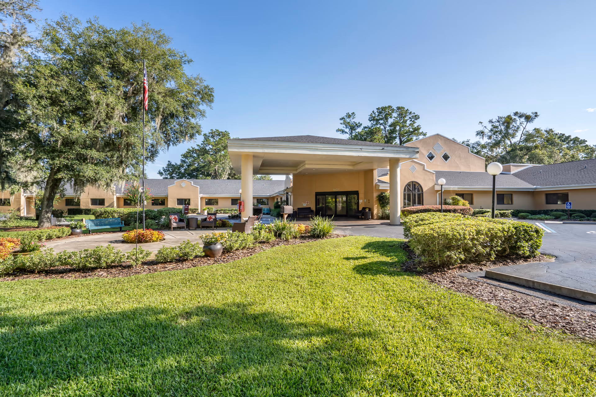 Exterior view of Brookdale Paddock Hills senior living facility showing the main entrance with a covered drop-off area, surrounded by well-maintained green lawns, shrubs, and trees under a clear blue sky.