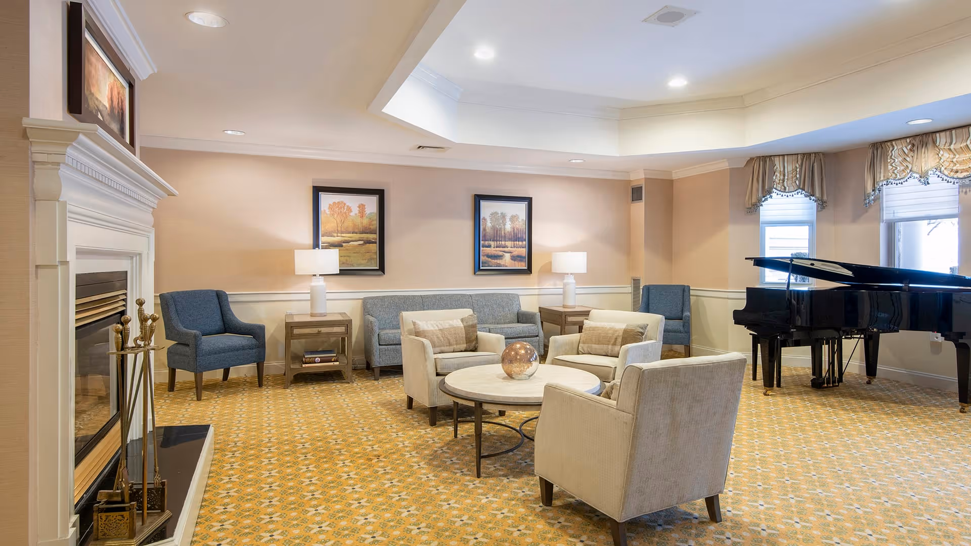 A cozy living room area with a fireplace on the left, a set of beige armchairs and a round coffee table in the center, a blue sofa and two blue armchairs against the back wall, two framed landscape paintings, two table lamps, and a black grand piano near two windows with decorative valances.