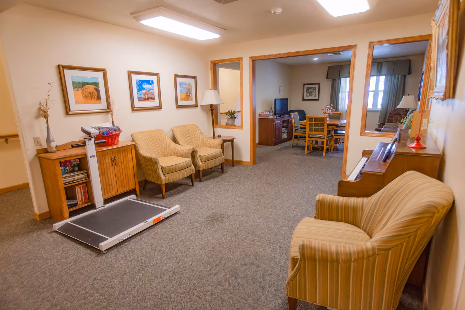 A cozy interior common area in an assisted living facility featuring beige walls and carpeted floors. The room has several striped upholstered chairs, a wooden cabinet with books and a basket on top, a floor lamp, framed landscape pictures on the wall, and a piano. Through an open doorway, another room is visible with a TV, wooden dining table, and chairs, along with windows letting in natural light.