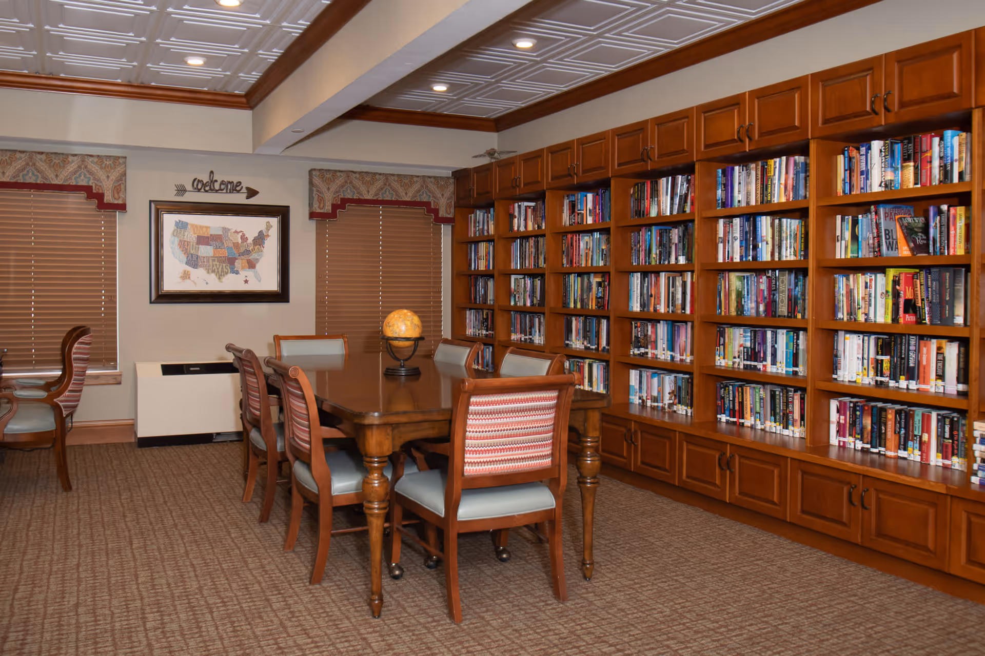 A cozy library room with wooden bookshelves filled with books along one wall. In the center, there is a wooden table surrounded by chairs with cushioned seats and backs. The room has carpeted flooring, two windows with closed blinds and decorative valances, and a framed map of the United States on the wall with a 'welcome' sign above it. The ceiling has recessed lighting and decorative molding.