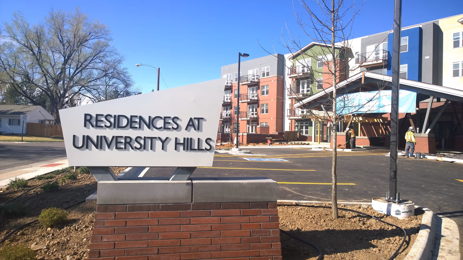 Outdoor view of the Residences at University Hills sign in front of a modern multi-story residential building with a parking lot and two workers in safety vests near the entrance.