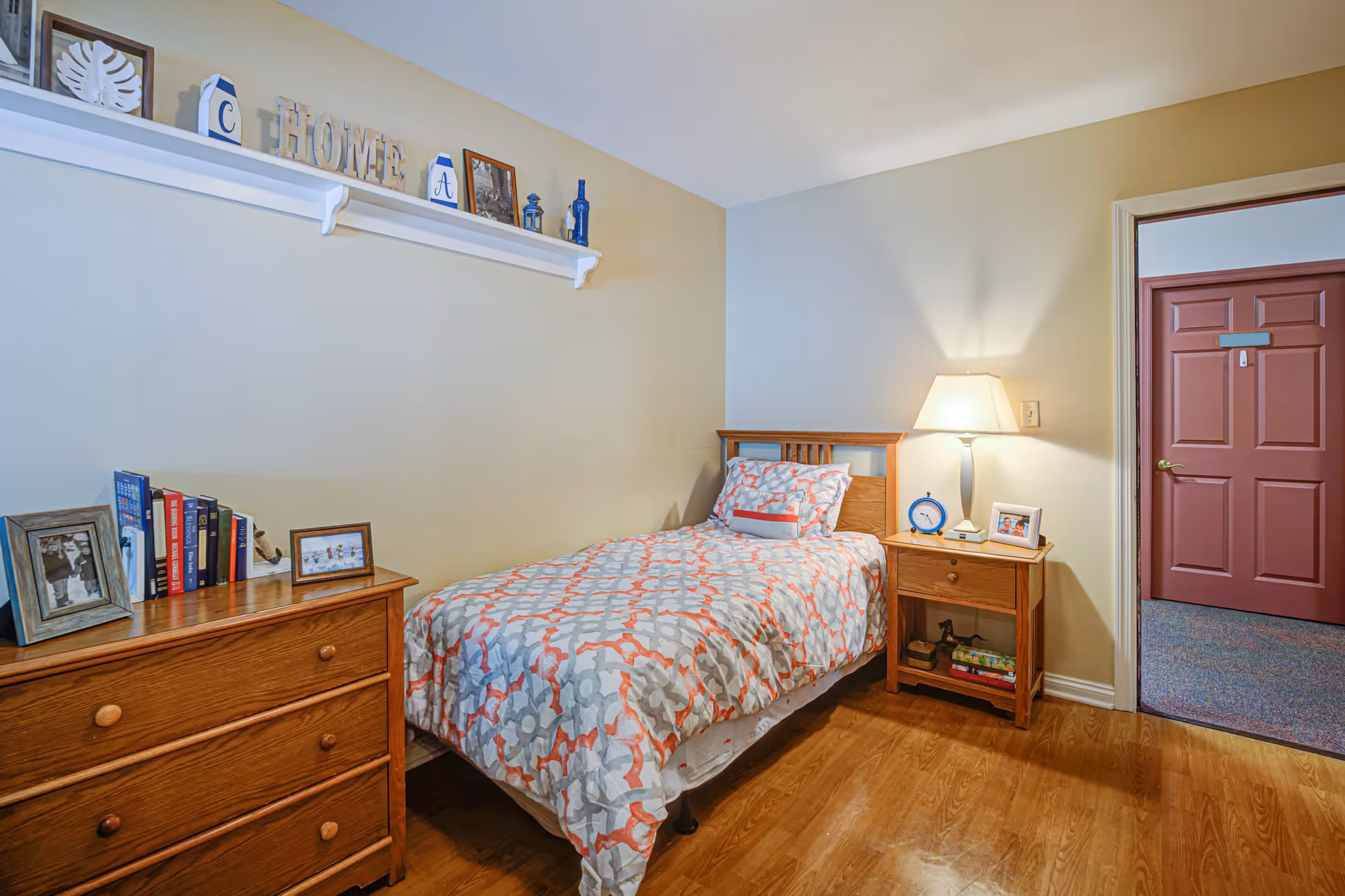 A small, cozy bedroom with a single bed covered in a patterned bedspread in shades of gray, white, and coral. Next to the bed is a wooden nightstand with a lamp, a blue alarm clock, and a framed photo. On the left side of the room is a wooden dresser with framed photos and books on top. Above the bed is a white wall shelf displaying decorative items including letters spelling 'HOME'. The room has light-colored walls and wooden flooring. A door with a reddish-brown color is open, showing a carpeted hallway outside.