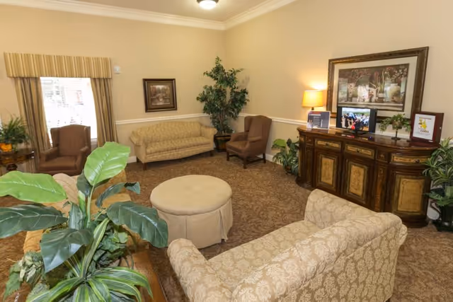 A cozy living room area with beige patterned sofas and armchairs arranged around a round ottoman. There is a wooden sideboard with a lamp, framed artwork, and a small TV on top. The room has beige walls, a window with curtains, and several green plants adding a touch of nature.