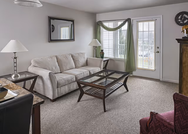 Living room with a beige sofa, glass-top coffee table, side tables with lamps, a window with green drapery, and a door to a balcony.