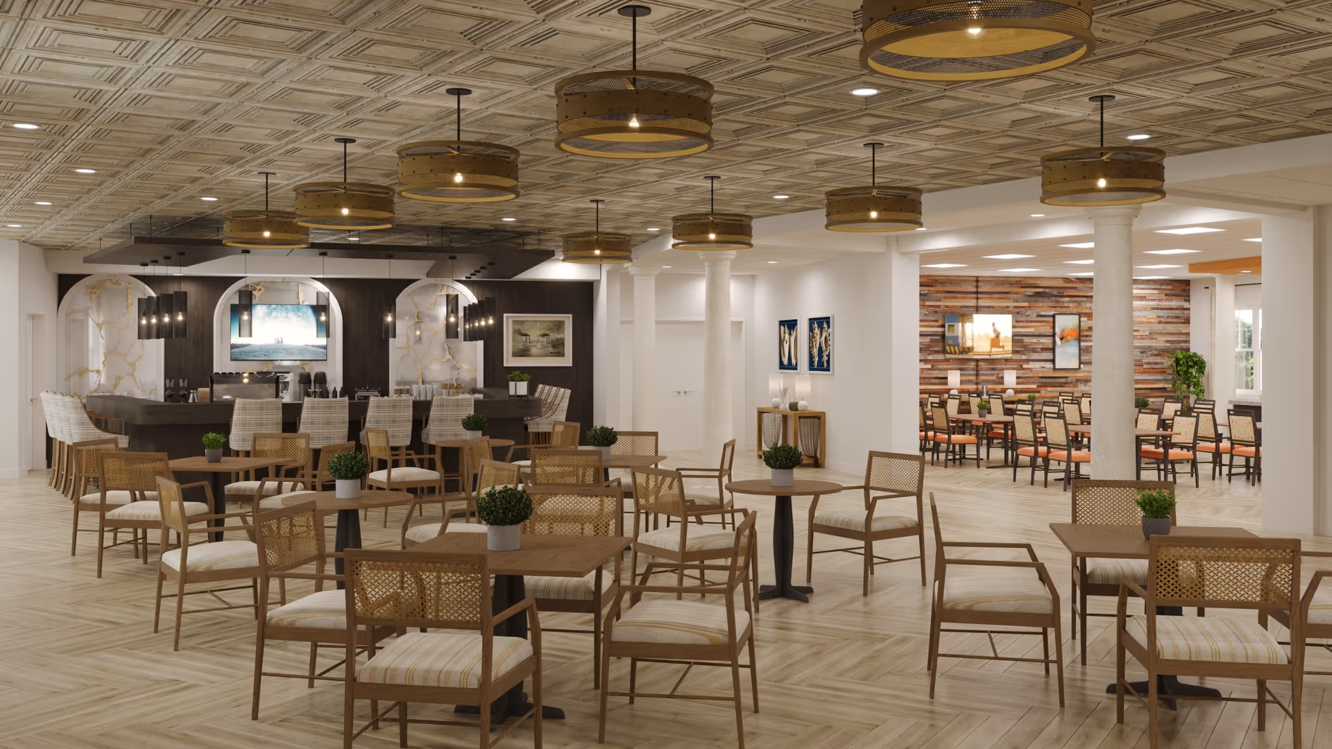 A spacious dining area with multiple wooden tables and chairs arranged neatly. The ceiling features decorative panels and several round pendant lights. In the background, there is a bar area with high chairs and a television mounted on the wall. The room has a light wood floor and white columns, with additional seating visible in an adjoining room with a wood-paneled accent wall and framed artwork.