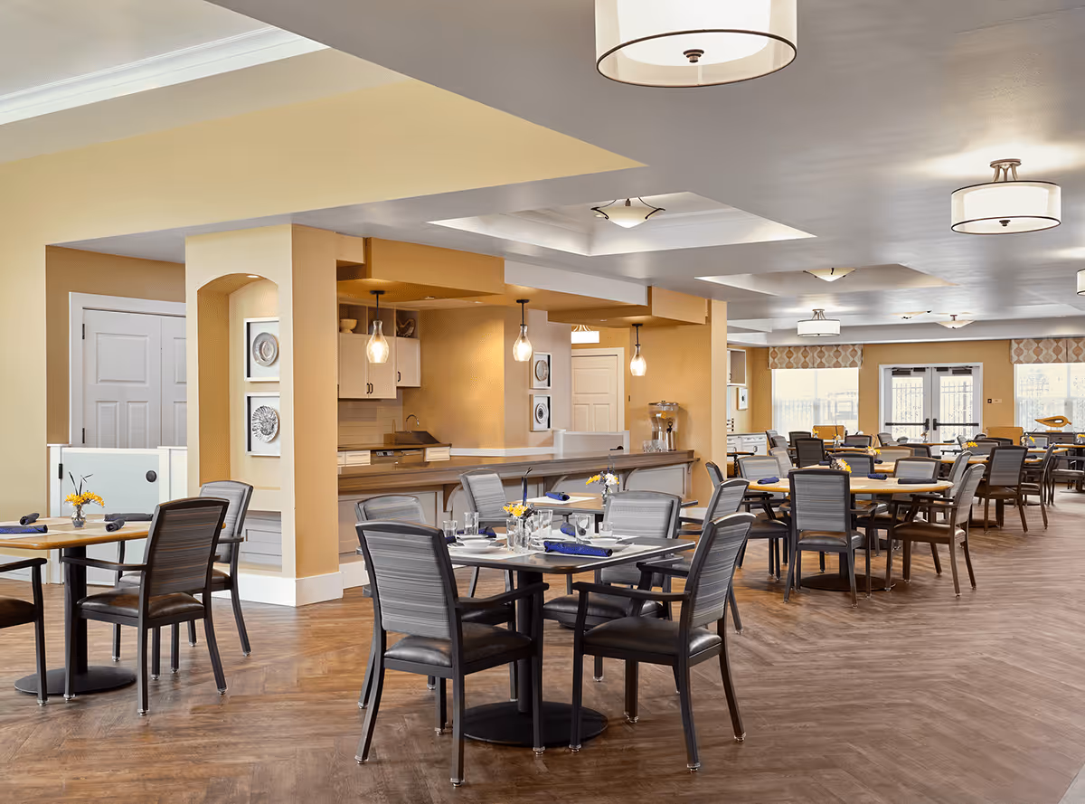 A spacious dining room in MorningStar Assisted Living & Memory Care of Boulder featuring multiple tables with chairs arranged neatly. The tables are set with plates, glasses, and napkins, and small flower arrangements add a decorative touch. The room has warm beige walls, pendant and ceiling lights, and large windows allowing natural light to enter.