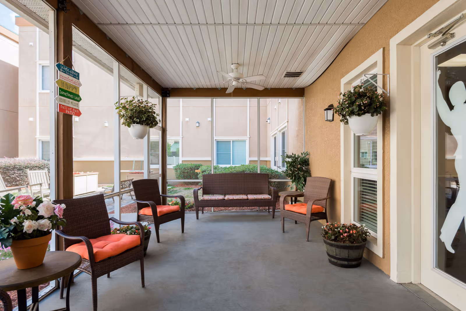 A covered outdoor patio area with wicker chairs and a loveseat, all with orange cushions. There are hanging flower pots and potted plants around the space. A ceiling fan is mounted on the white paneled ceiling. The patio is enclosed with screen walls, and the exterior of the building is visible in the background. A colorful wooden sign hangs on a post with welcoming messages.