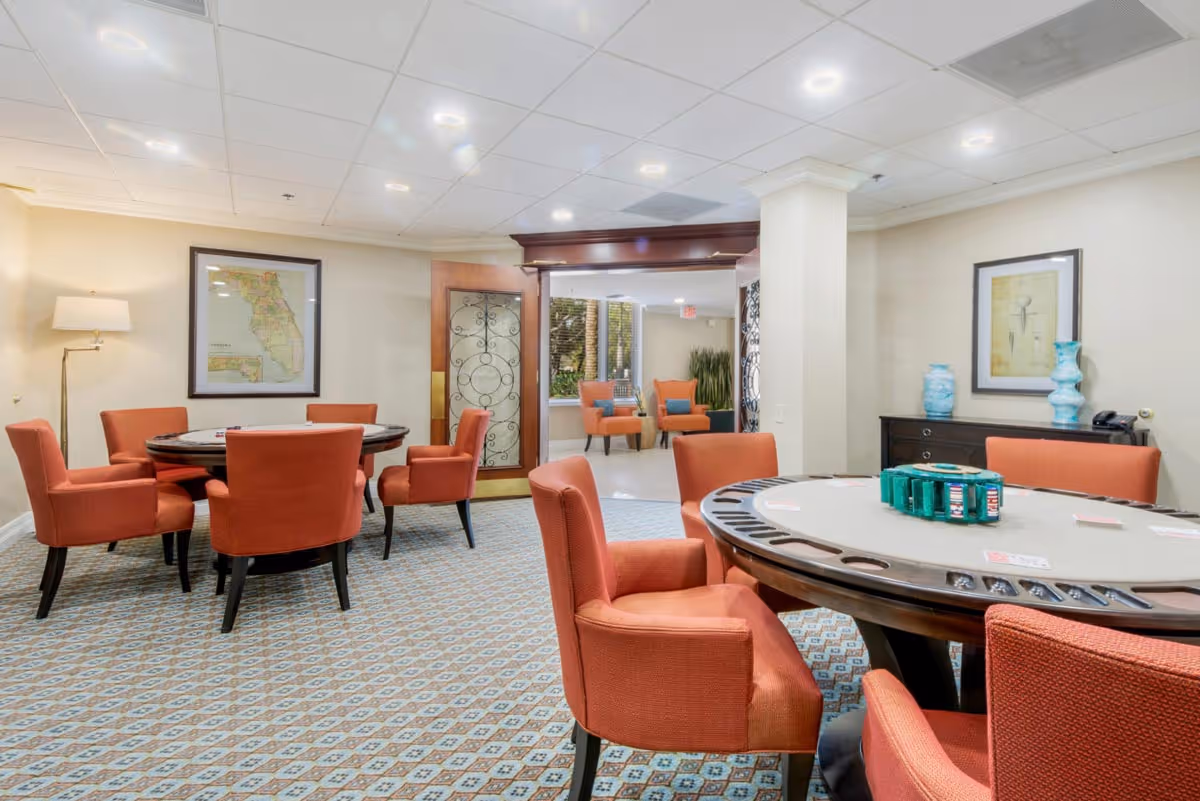 Well-lit communal game room with round card tables surrounded by orange upholstered chairs.