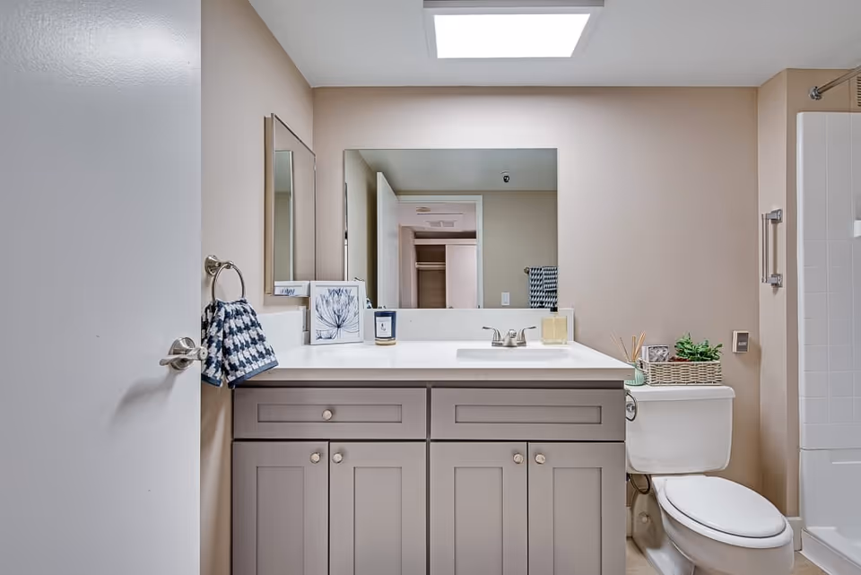 A clean and modern bathroom featuring a light gray vanity with a white countertop, a rectangular mirror above the sink, a toilet with a basket of decorative items on top, a towel hanging on a ring beside the vanity, and a shower area with white tiles visible on the right side.