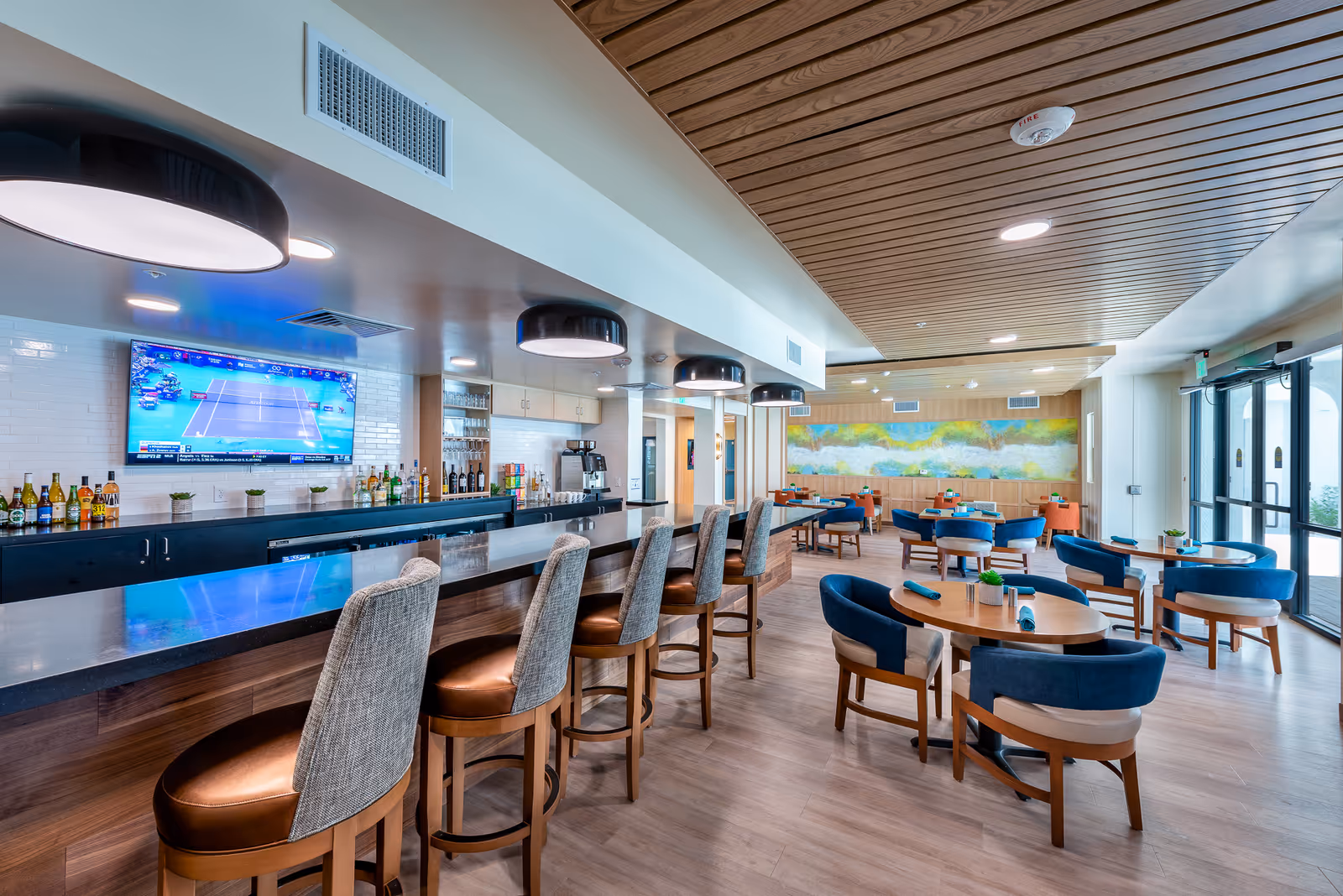 A modern dining area with a long bar counter featuring high chairs with cushioned seats and backs. Behind the bar, there are bottles of beverages and a large flat-screen TV mounted on a white tiled wall. The dining area has several round wooden tables with blue cushioned chairs, each table set with rolled napkins. The ceiling has wooden paneling and recessed lighting, and large windows allow natural light to enter the space.