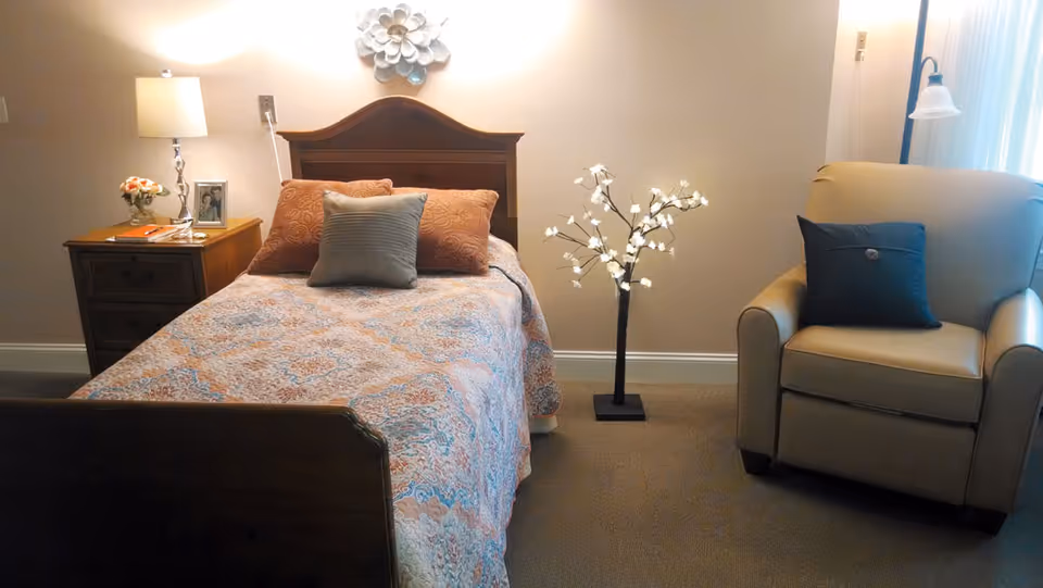 A cozy bedroom featuring a single bed with a patterned bedspread and three pillows, a wooden nightstand with a lamp, a small flower vase, and a framed photo. Next to the bed is a decorative floor lamp shaped like a tree with white flowers. To the right is a beige armchair with a dark blue cushion, positioned near a window with sheer curtains.
