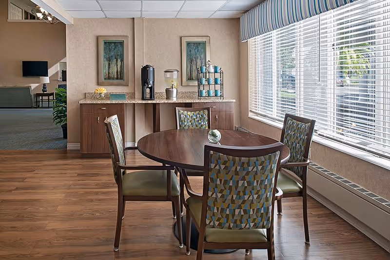A dining area in a senior living facility with a round wooden table surrounded by four chairs with patterned upholstery. Behind the table is a countertop with a coffee dispenser, a water dispenser, and a rack holding blue cups. Two framed paintings hang on the wall above the countertop. Large windows with blinds allow natural light to fill the room.