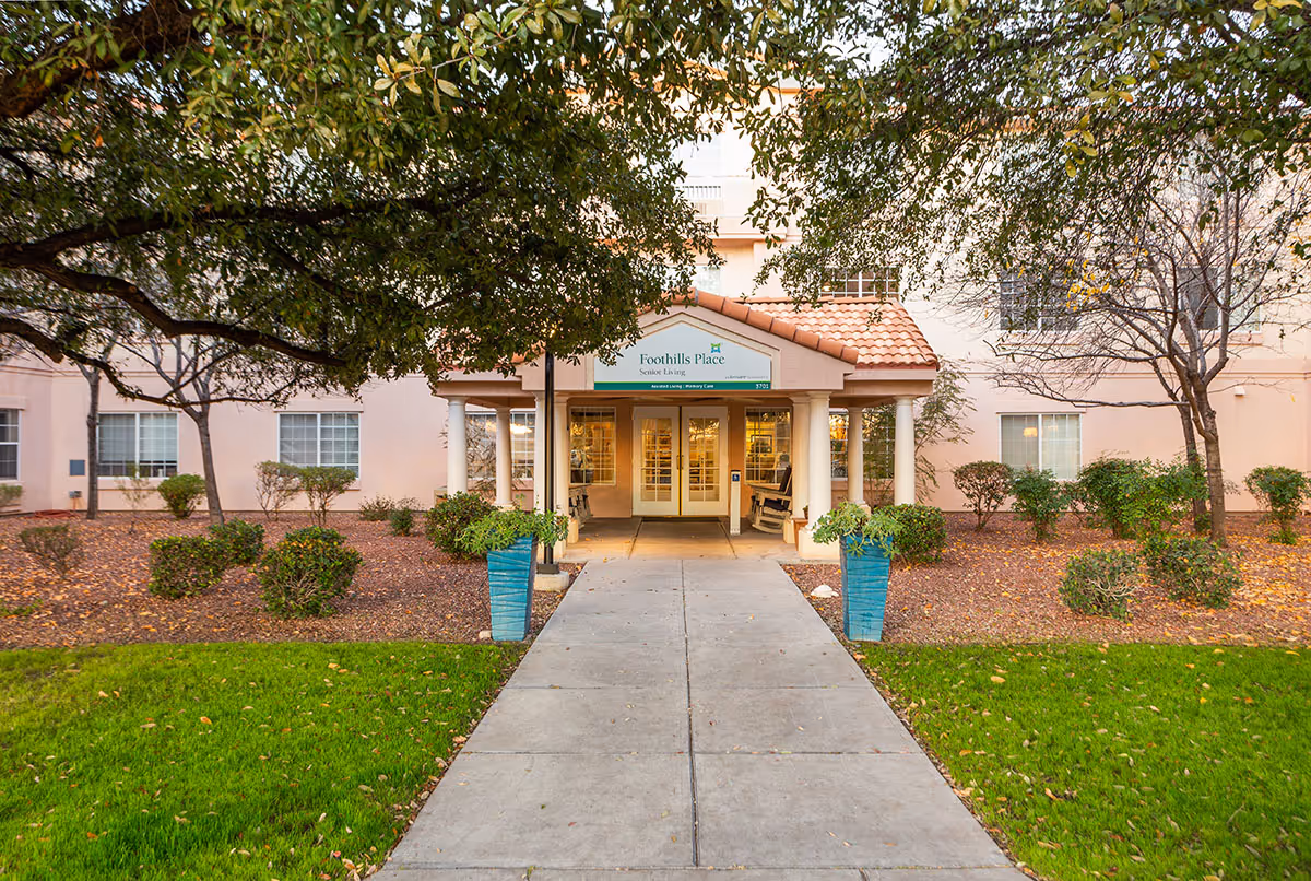Front entrance of a senior living facility named Foothills Place Senior Living, with a covered walkway, two blue planters with greenery, and surrounding landscaping including grass, bushes, and trees.