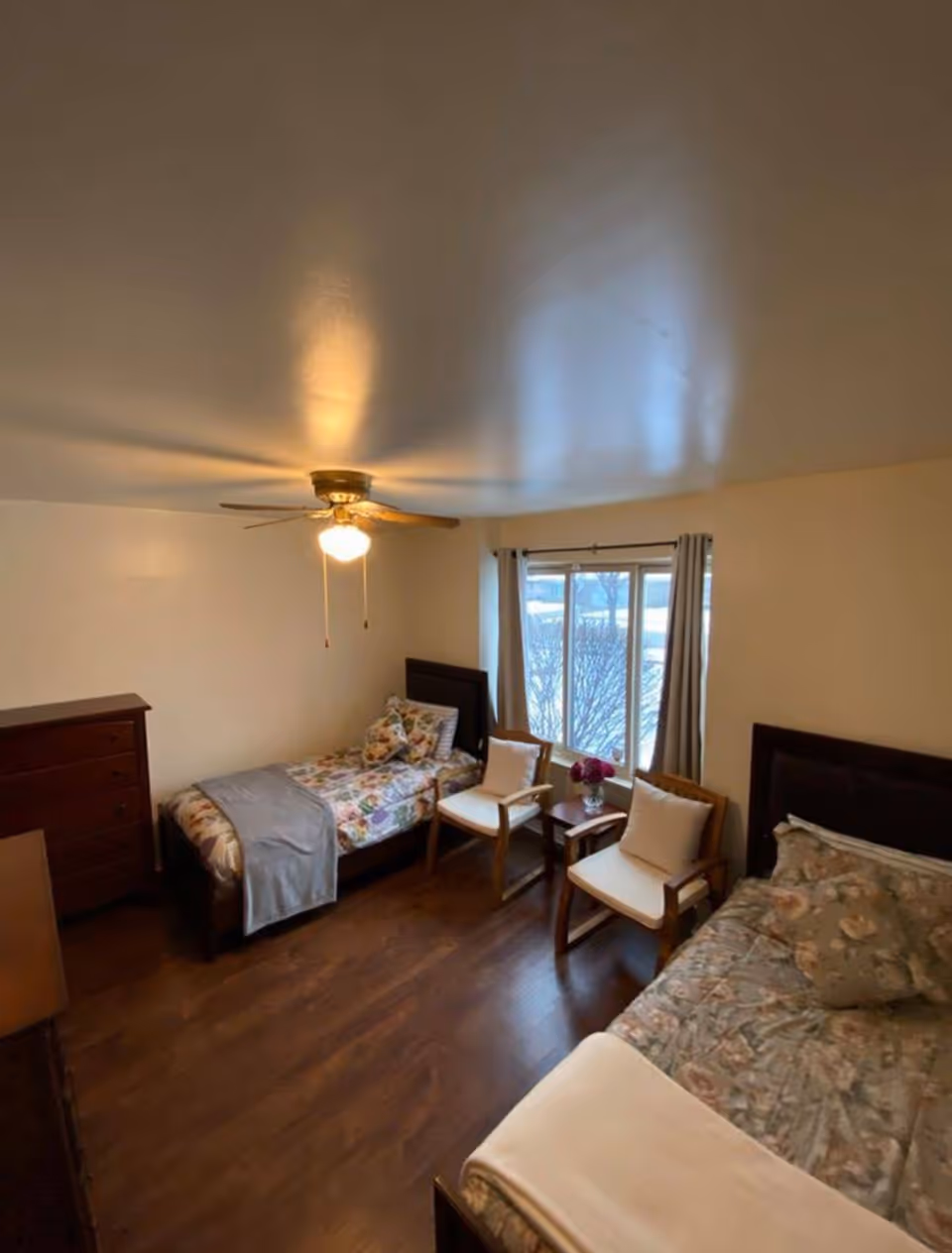 Well-lit bedroom with two twin beds, a ceiling fan, two chairs and a small table by a window, and wood flooring.