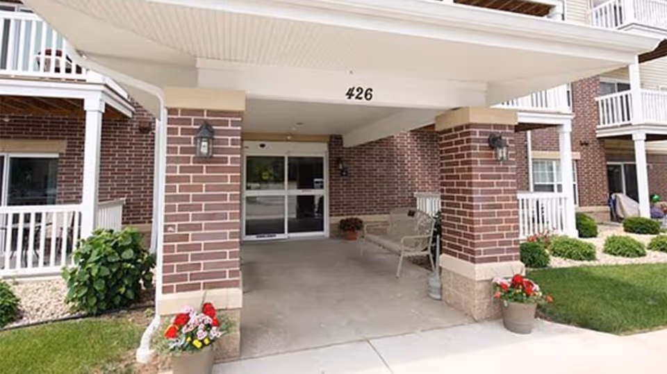 Entrance to a senior apartment building with brick pillars, a covered walkway, potted flowers, and a bench near the glass doors. The building number 426 is displayed above the entrance.