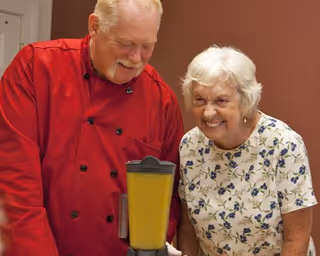 An elderly man in a red chef coat and an elderly woman in a white floral shirt smiling together in a kitchen setting with a blender containing a yellow mixture in front of them.