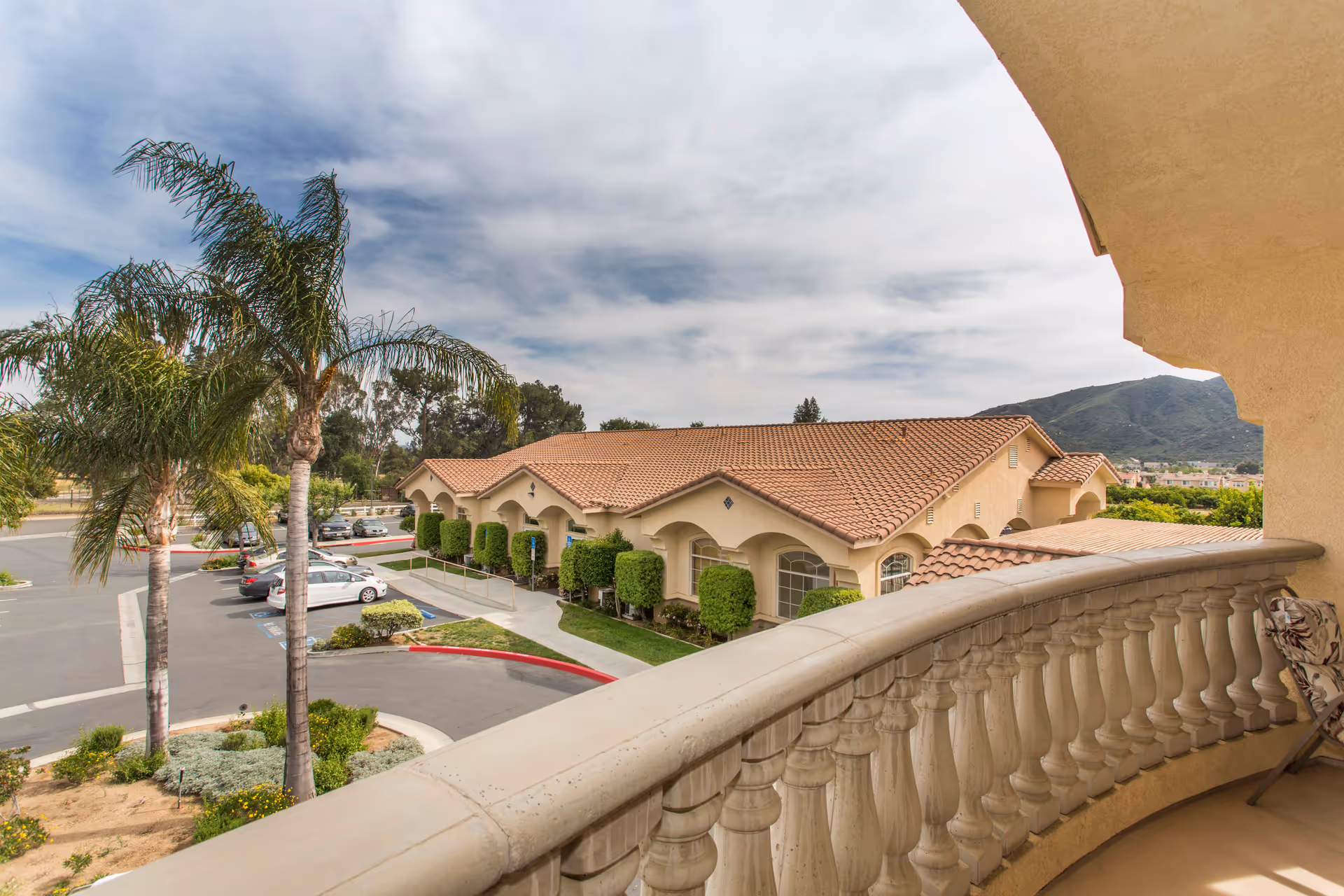 Balcony view overlooking a Mediterranean-style senior living building with red tile roofs, palm trees and a parking lot.