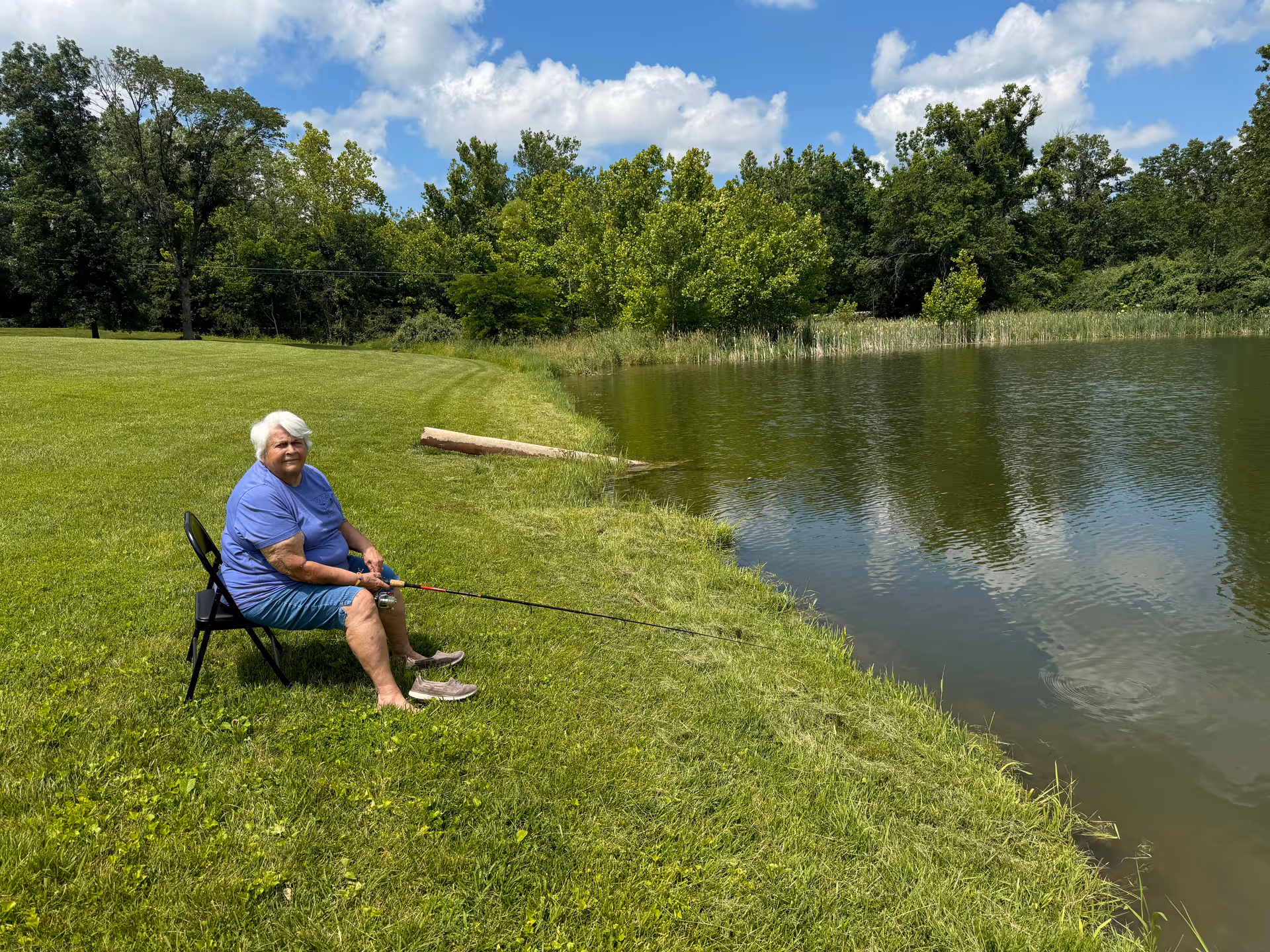 An elderly woman with white hair sitting on a black folding chair on a grassy bank, fishing in a calm pond surrounded by green trees under a partly cloudy blue sky.