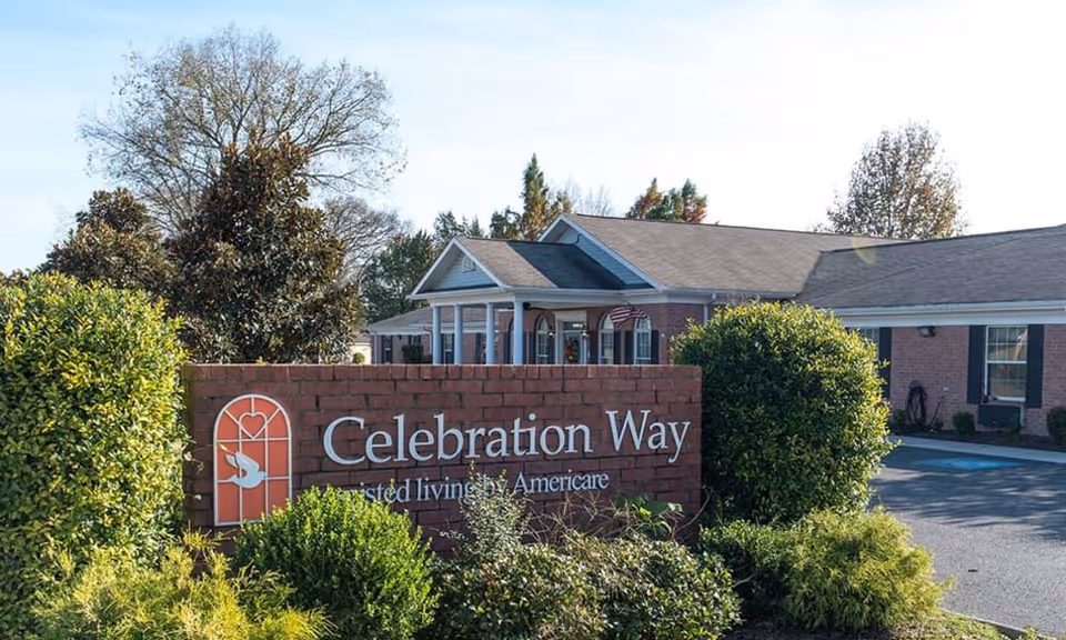 Exterior view of Celebration Way assisted living facility showing a brick sign with the facility name surrounded by bushes and trees, with the building and an American flag in the background under a clear sky.