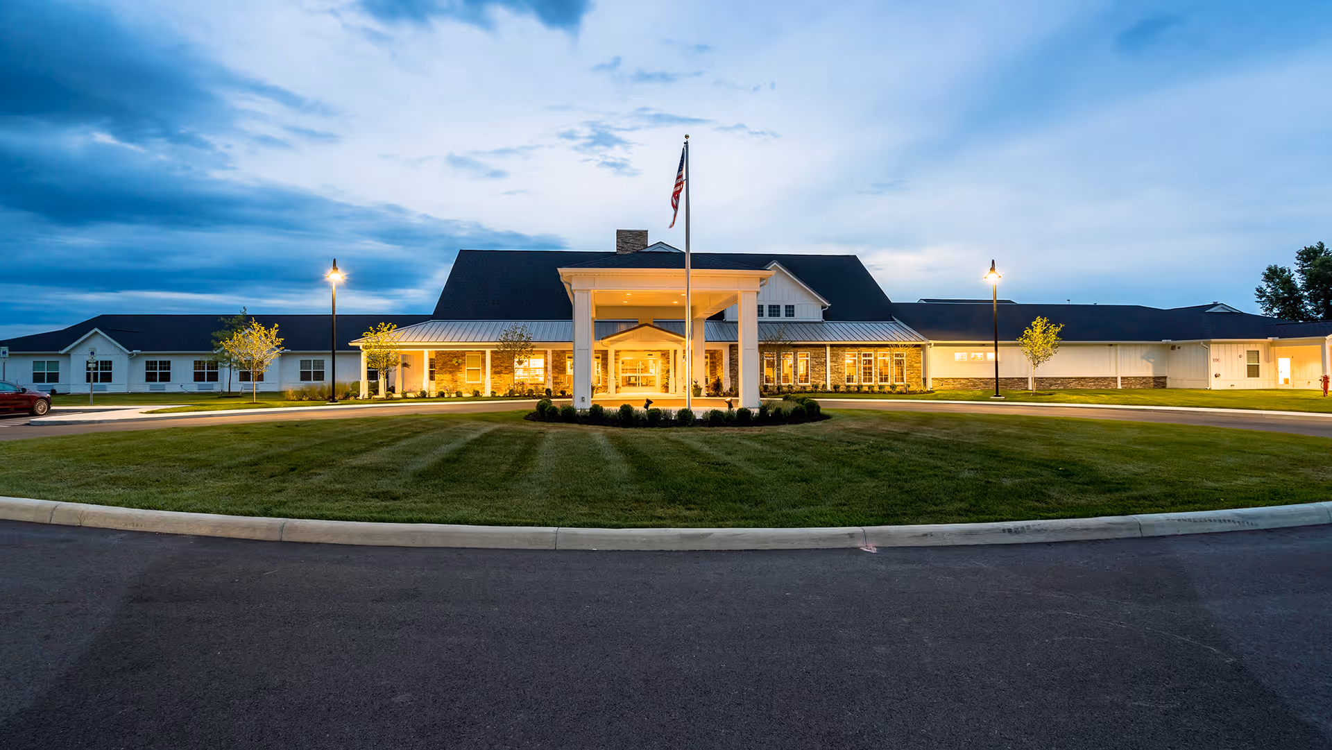 Front exterior view of Walnut Crossing facility at dusk with a circular driveway, well-maintained lawn, flagpole with an American flag, and warm lights illuminating the entrance.