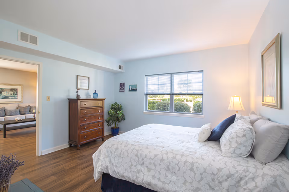 A bright bedroom with a large bed covered in white floral bedding and multiple pillows. A wooden chest of drawers stands against the left wall with decorative items on top. A window with blinds lets in natural light, and a framed picture hangs on the right wall. The room has light blue walls and wood flooring. Through an open doorway, a living room with a sofa and coffee table is visible.