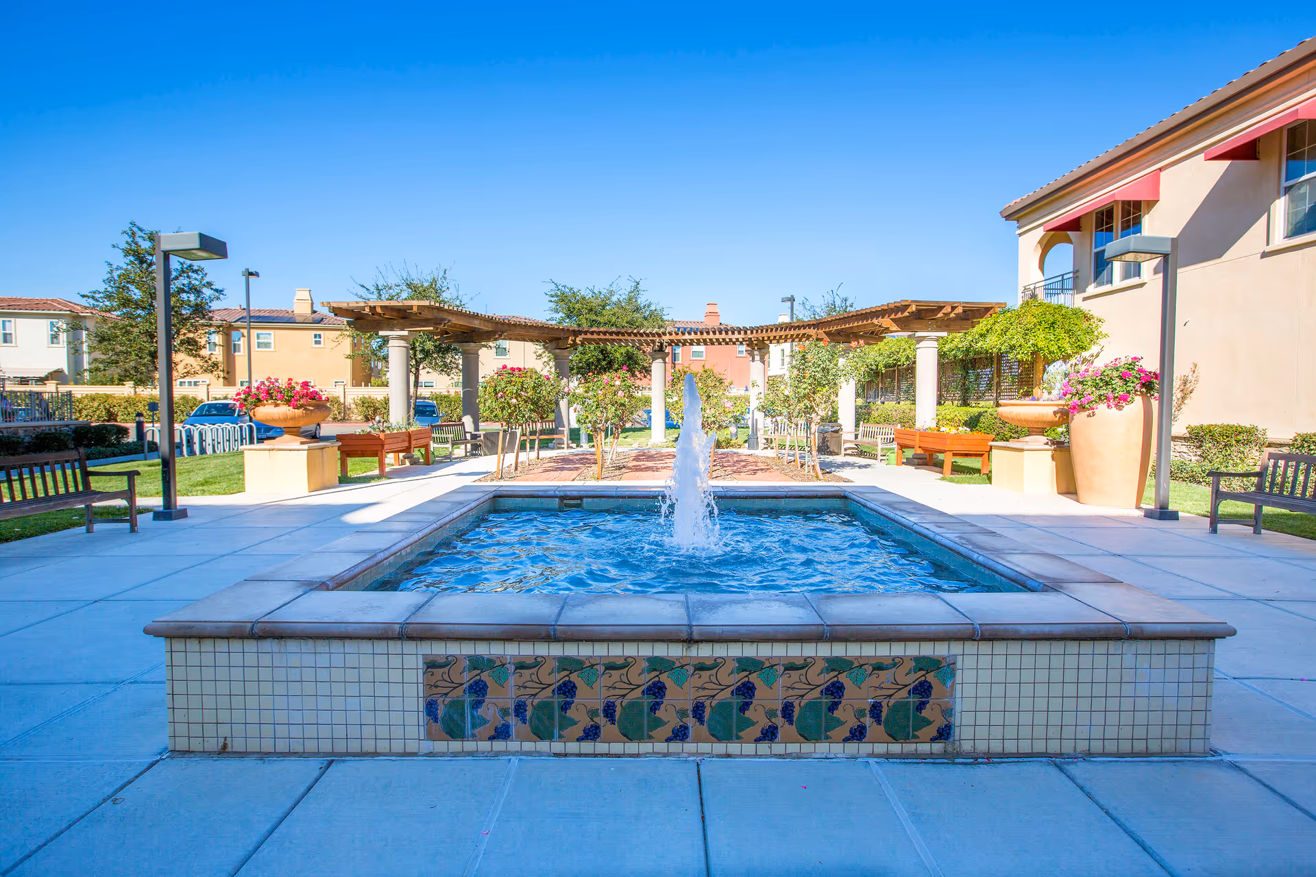 Sunny outdoor courtyard with a central decorative fountain, benches, pergolas and potted plants in front of residential buildings.