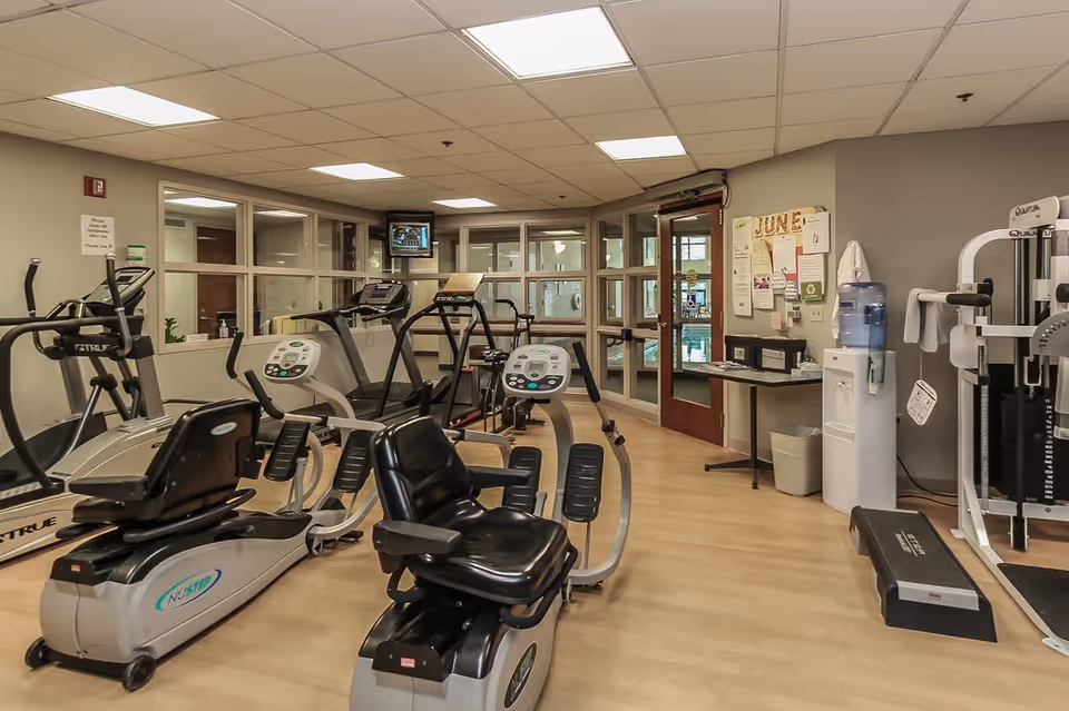 Indoor fitness room with exercise equipment including recumbent bikes, treadmills, elliptical machines, and a multi-station gym machine. The room has light wood flooring, beige walls, a water cooler, and a bulletin board with notices. There is a door with glass panels leading to another area.