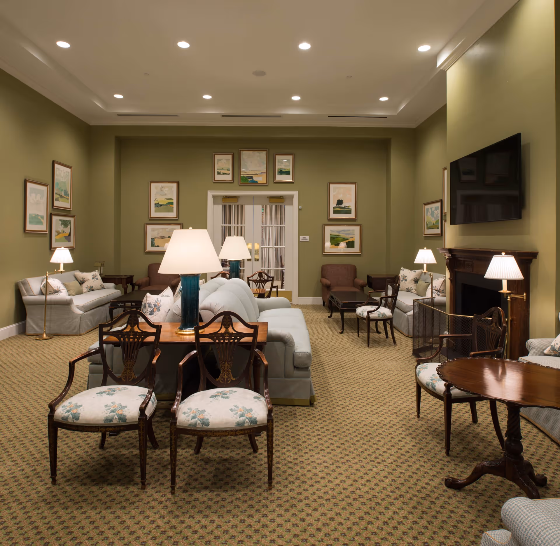 A spacious living room with green walls and patterned carpet. The room is furnished with multiple sofas, armchairs, and wooden chairs with floral upholstery. Several table lamps with white shades are placed on wooden tables. A fireplace with a dark wooden mantel is on the right wall, above which is a mounted flat-screen TV. The walls are decorated with framed landscape paintings. Double glass doors are visible at the far end of the room.